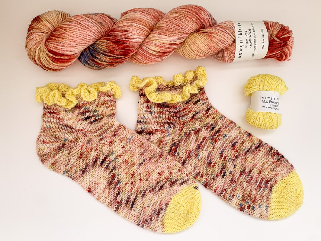 A skein of Cowgirlblues Proper Sock yarn, colour Bridget's Blush, lies horizontally across the top, beneath is a pair of hand knit Midnight Dancer socks and a mini ball of Proper Sock in Lemon