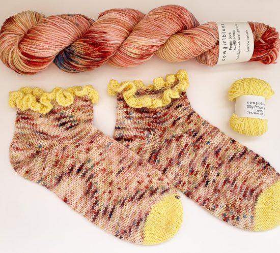 A skein of Cowgirlblues Proper Sock yarn, colour Bridget's Blush, lies horizontally across the top, beneath is a pair of hand knit Midnight Dancer socks and a mini ball of Proper Sock in Lemon