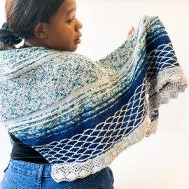 Woman looks backwards over right shoulder and has a hand knit shawl draped across her back