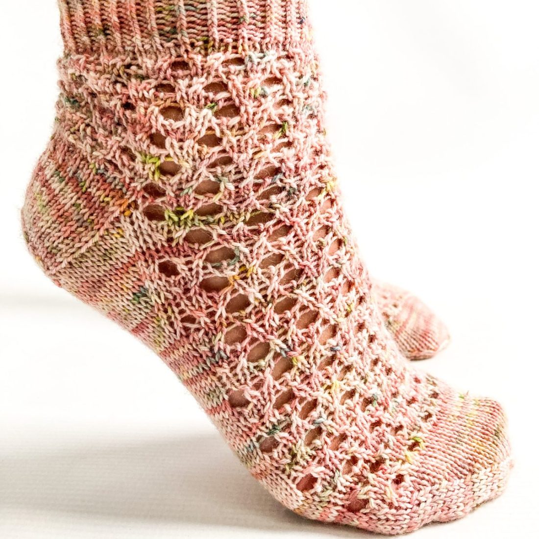 Foot on tiptoe wearing a a hand knit sock in a lace pattern and peachy pink variegated sock yarn
